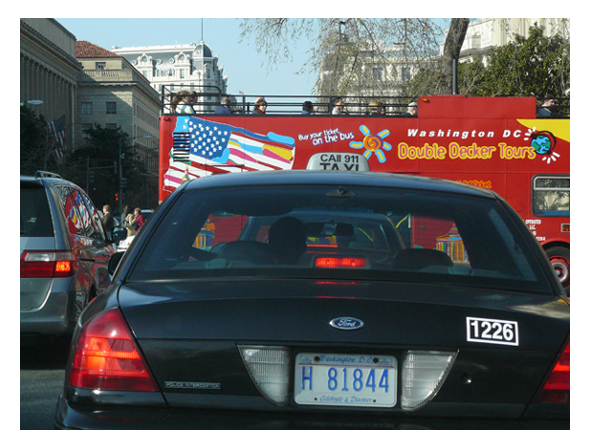 A Ford Crown Victoria taxi in traffic, facing a colorful, double-decker sightseeing bus.